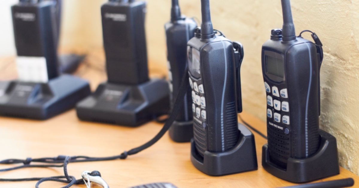 Several black two-way radios with white buttons and digital displays sitting in charging docks on a wooden table.