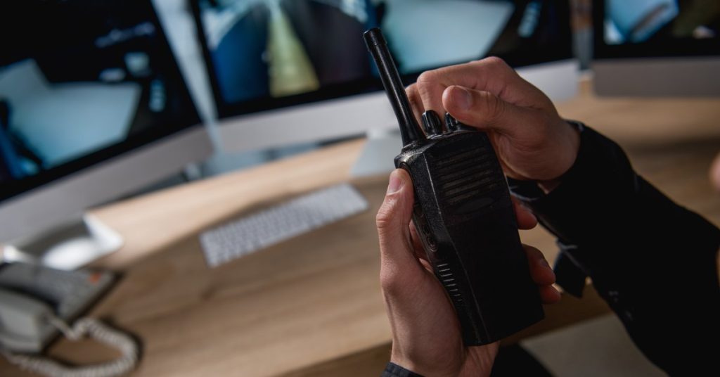 An individual twisting a knob on the top of a black two-way radio. There are several computer monitors in the background.