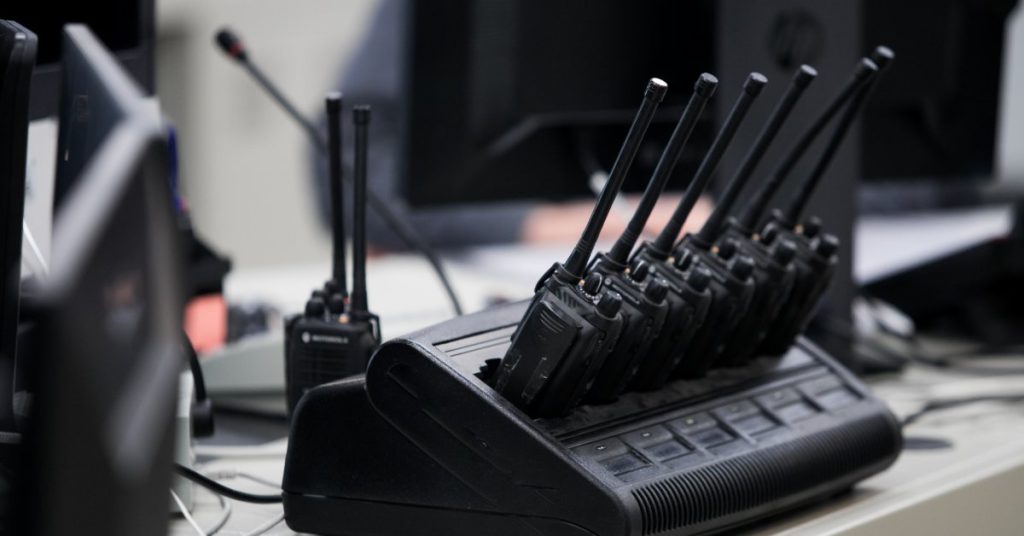 Several black two-way radios with antennas sitting in a large charging dock. There is a computer monitor in the background.