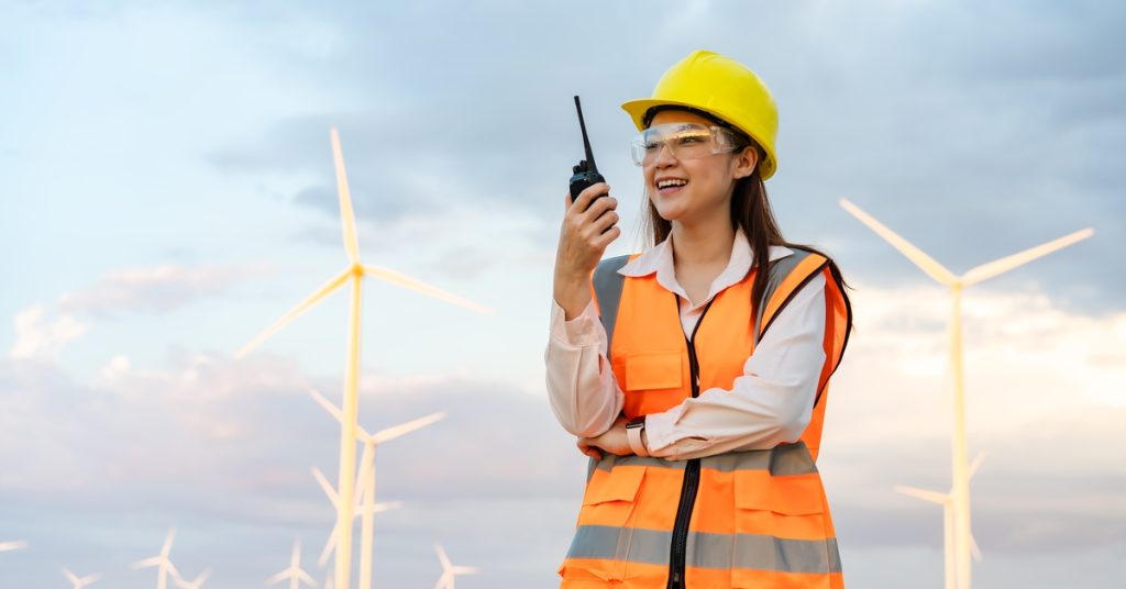 A woman in an orange safety vest, yellow hard hat, and safety glasses using a two-way radio in front of wind turbines.