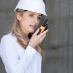 A blonde-haired woman in a white collared shirt and white hard hat speaking into an orange two-way radio.