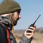 A man in a black and gray jacket and green beanie using a black two-way radio while standing outside.