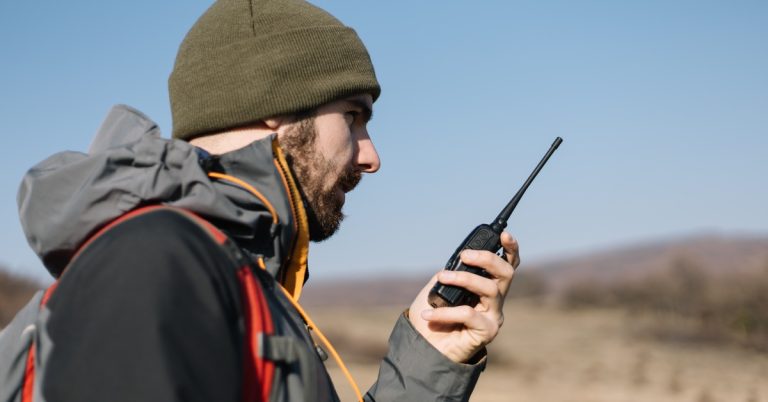A man in a black and gray jacket and green beanie using a black two-way radio while standing outside.