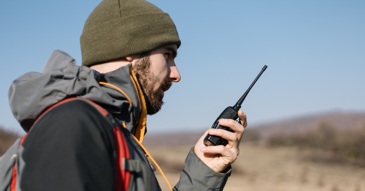 A man in a black and gray jacket and green beanie using a black two-way radio while standing outside.
