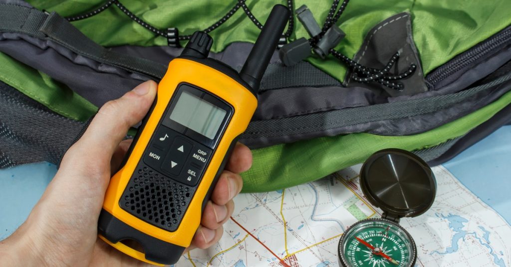 A close-up of an individual holding a yellow and black two-way radio in front of a map, a compass, and a green backpack.