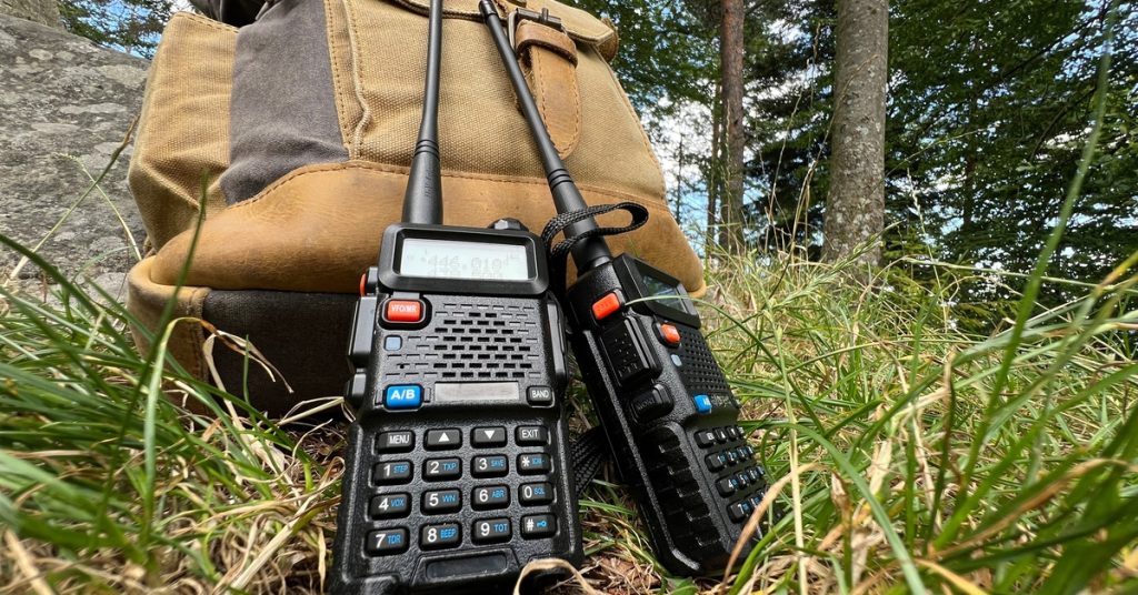 A close-up of two black two-way radios resting against a brown backpack in the grass. There are trees in the background.