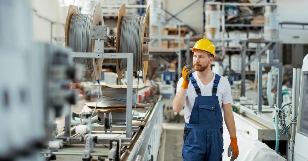 A man in blue overalls, a yellow hard hat, and orange gloves using a two-way radio in a factory with large equipment.