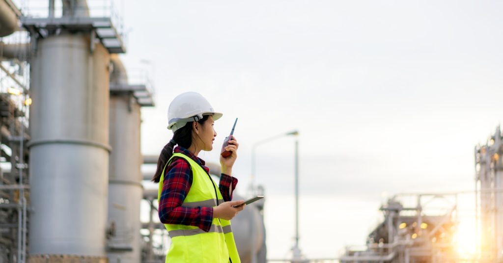 A woman in a white hard hat and yellow safety vest using a two-way radio while standing outside an oil refinery.