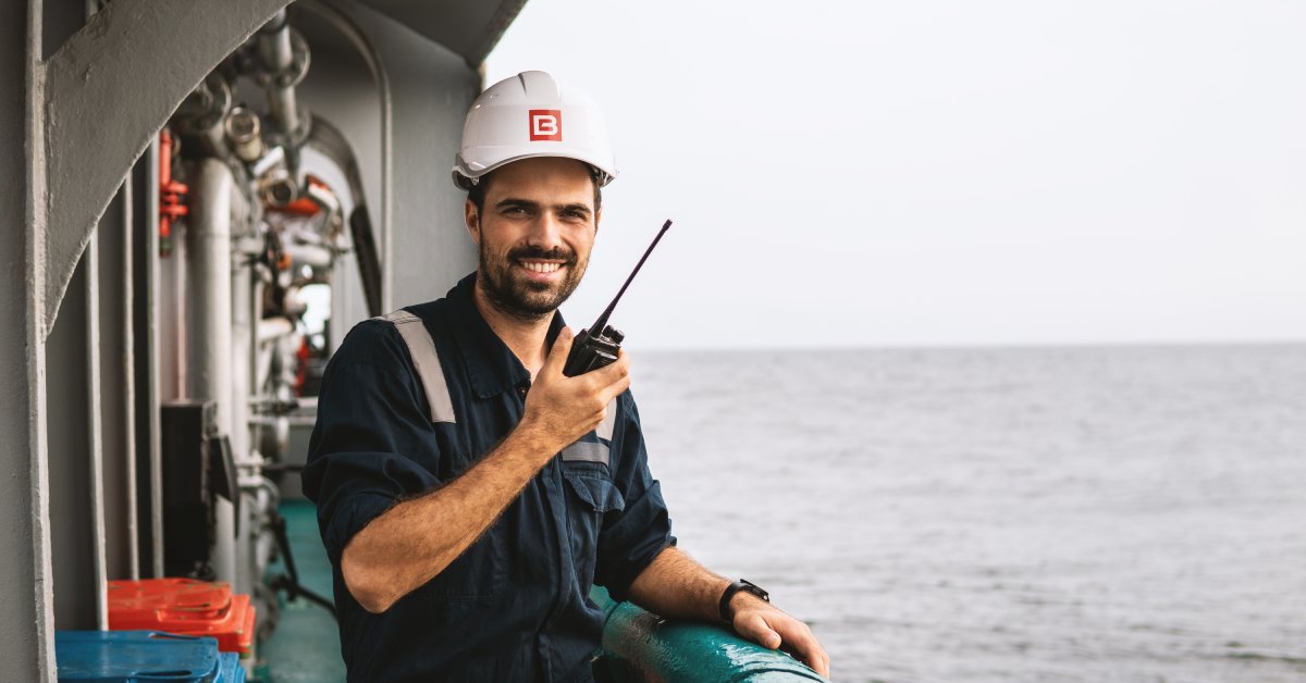 A smiling man in a black jumpsuit and white hard hat using a two-way radio while standing on a boat deck.