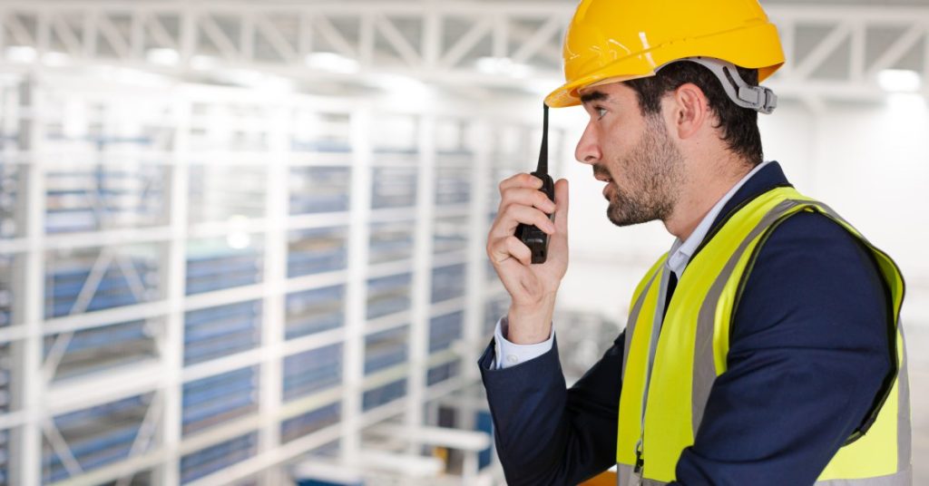 How Radio Signal Strength Impacts Two-Way Radio Batteries A man in a blue suit, yellow safety vest, and yellow hard hat using a black two-way radio in a large warehouse.