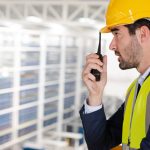 A man in a blue suit, yellow safety vest, and yellow hard hat using a black two-way radio in a large warehouse.