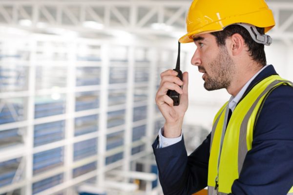 A man in a blue suit, yellow safety vest, and yellow hard hat using a black two-way radio in a large warehouse.