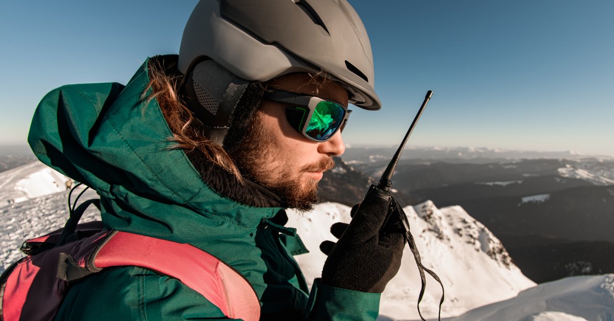 A man wearing a helmet, jacket, gloves, sunglasses, and a backpack while using a two-way radio on top of a snowy mountain.