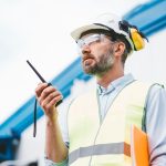 A man in a safety vest, hard hat, earmuffs, and safety goggles holding an orange folder under his arm while using a radio.