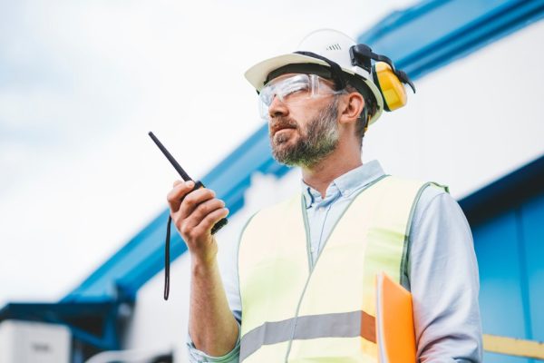 How To Keep Your Two-Way Radio Operating at Peak Levels A man in a safety vest, hard hat, earmuffs, and safety goggles holding an orange folder under his arm while using a radio.