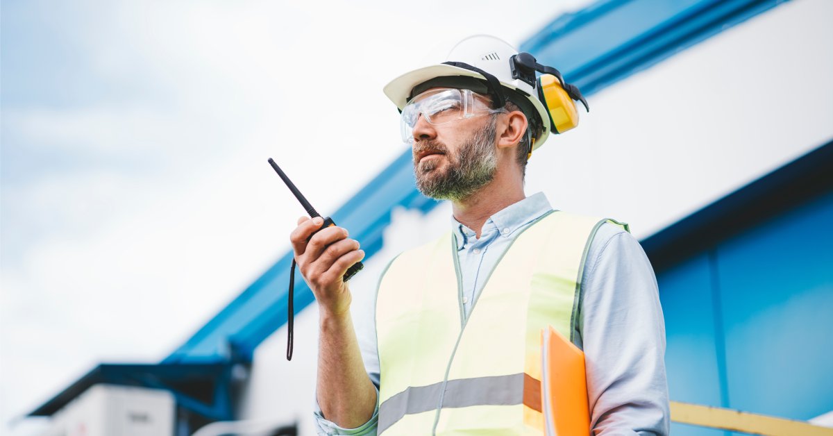 A man in a safety vest, hard hat, earmuffs, and safety goggles holding an orange folder under his arm while using a radio.