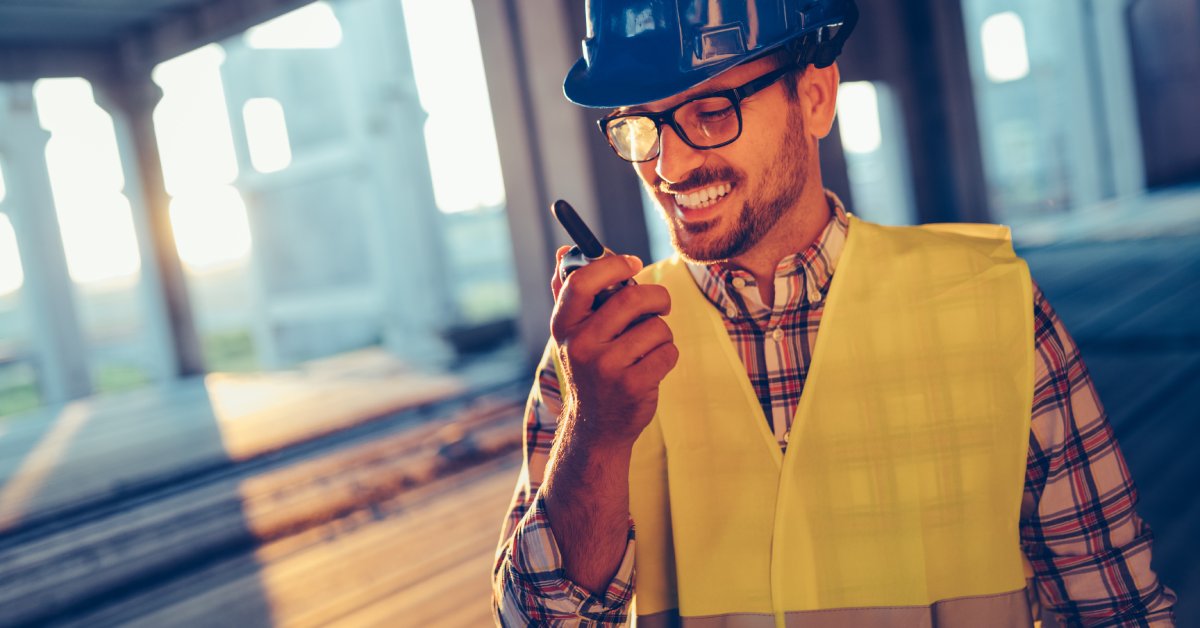 A smiling man in a plaid shirt, yellow safety vest, and blue hard hat using a two-way radio at a construction site.